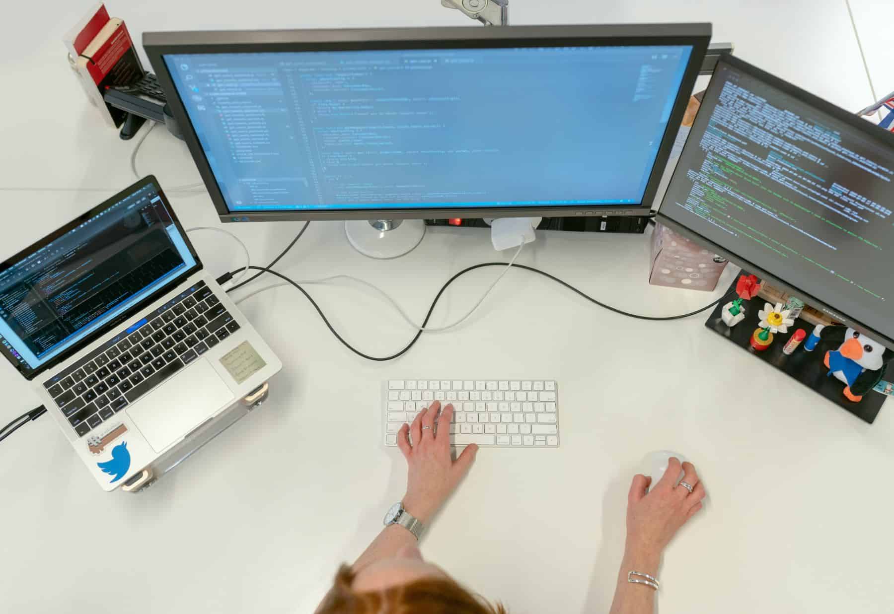 This image features a laptop, computer monitors, a computer keyboard, a person. Additional details include: electronic device. The image has dominant colors like cyan, grey, and white.